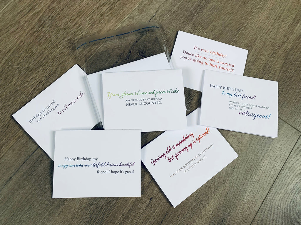 Six white birthday-themed notecards are overlapping on a wooden background. Each card has a mix of block and script font in an array of colors. Birthday Fun Notecards by Stationare.