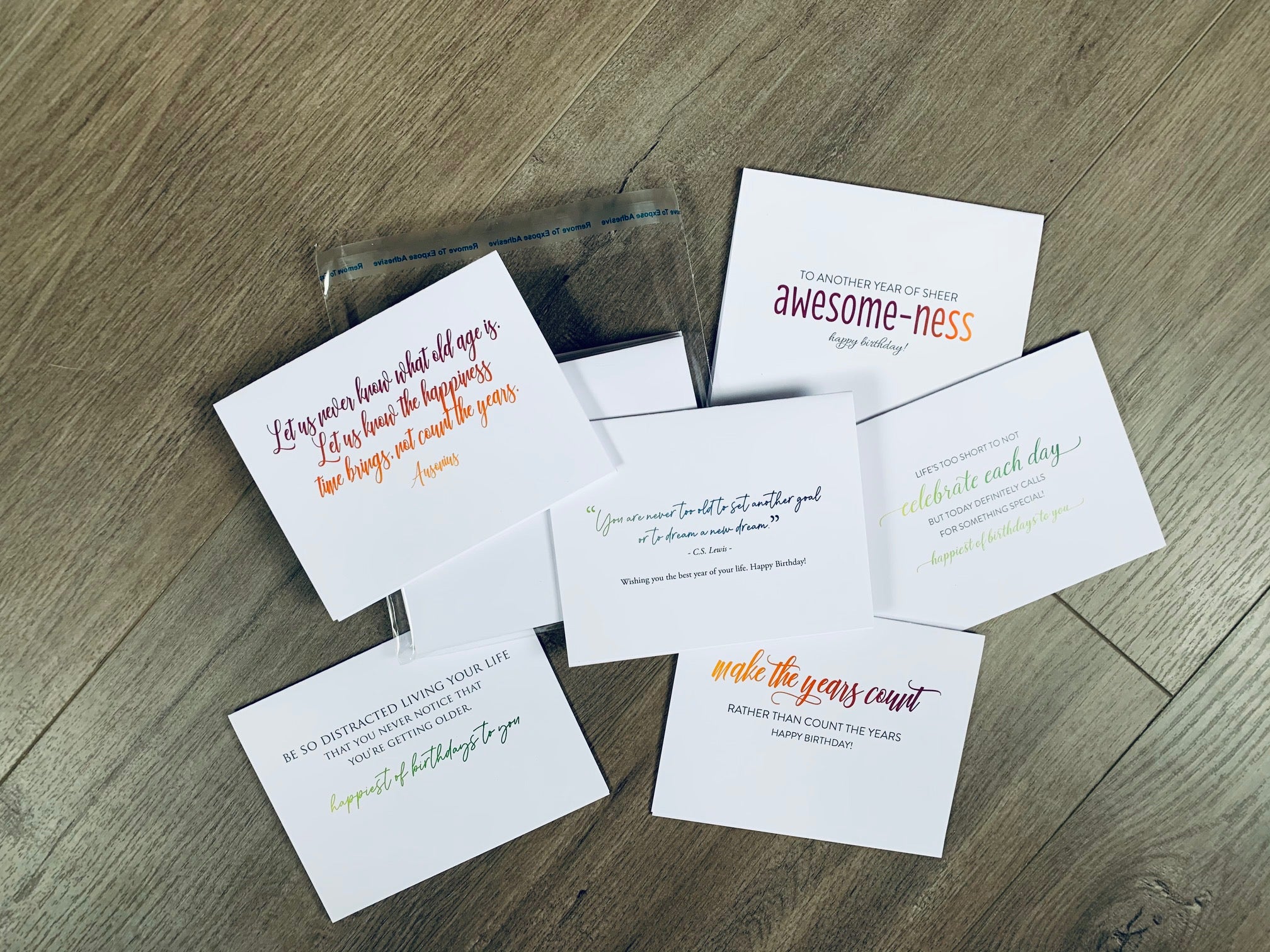 Six white birthday cards are overlapping on a wooden background. Each card is a mix of block and colorful script prints. The sayings are all encouraging, inspiring quotes about celebrating birthdays. Birthday Inspirations by Stationare.