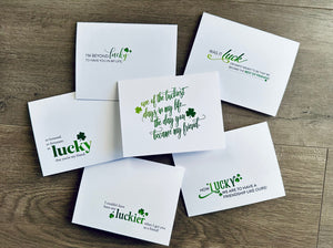 Six white Irish-themed notecards lie on a wooden background. Each card has a mix of fonts in differing greens and share a friendship-themed saying about luck.