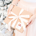 A female wearing a pale pink skirt is holding a light brown box with white polka dots and a white bow. In the bottom right hand  corner of the box is a white rectangular sticker tag that says "Merry Christmas" in a red script. Merry Christmas Sticker Gift Tag by Stationare.