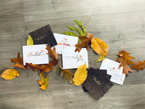 Four thanksgiving-themed notecards lie on a gray wooden floor surrounded by colorful fall leaves. On the top left and bottom right corner are cocktail napkins that read "thankful" and "thankful for cheese & wine"