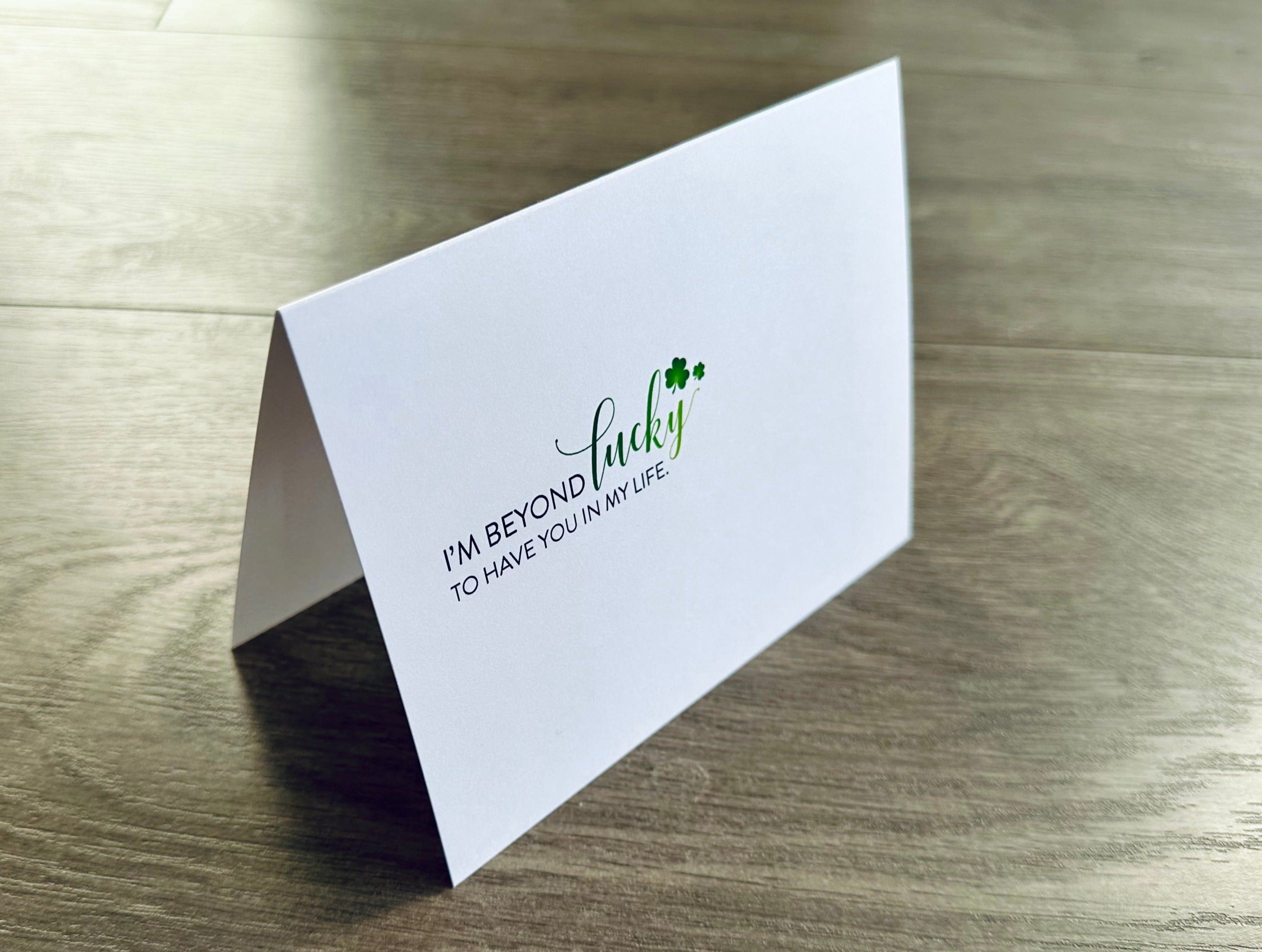 A folded white notecard says "I'm beyond lucky to have you in my life" on the front with two small green shamrocks. Friendship Luck by Stationare.