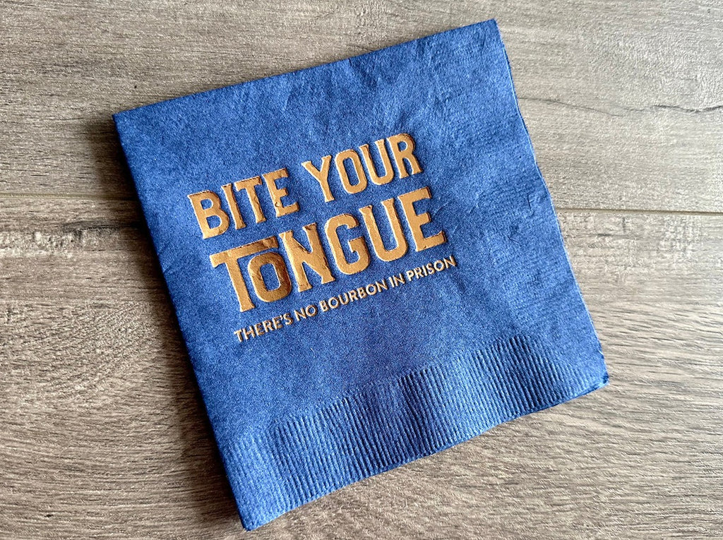 A blue cocktail napkin lies on a gray wooden background. In the center of the napkin is metallic gold print that reads, " Bite your tongue. There's no bourbon in prison."