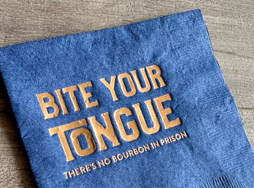 Closeup of a blue napkin on a gray wood background, showing the bold metallic gold print that reads: "Bite your tongue. There's no bourbon in prison."