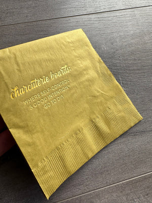 An angled image of a golden yellow cocktail napkin. In gold foil printing, it reads: charcuterie boards: Where self-control and good intentions go to die. By Stationare