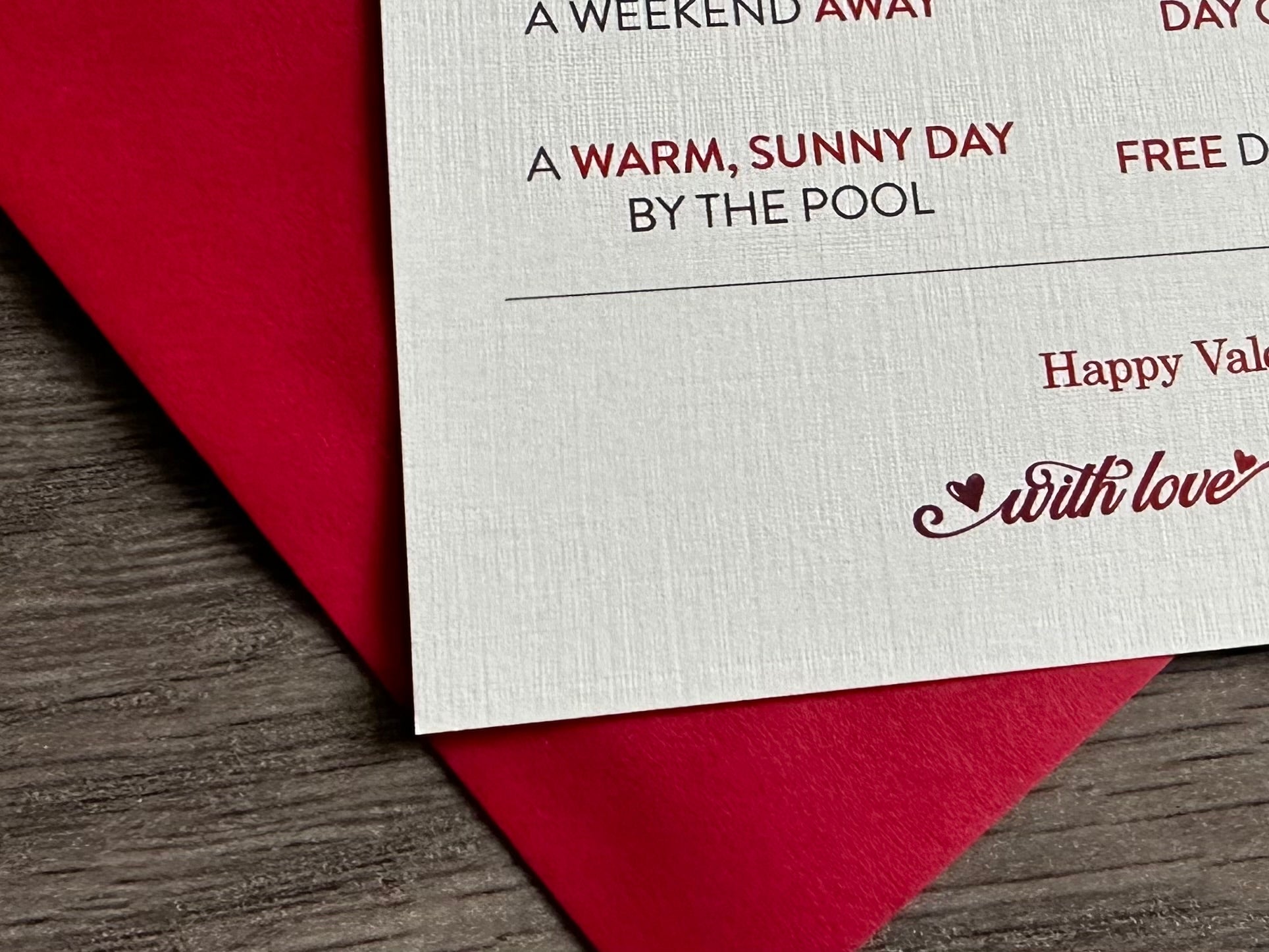 A closeup photo of an off-white card with block printing of "a warm, sunny day by the pool" lies on top of a red envelope and wood background. Portions of other sayings are shown, and at the bottom of the card, it reads "with love." Valentine card by Stationare.