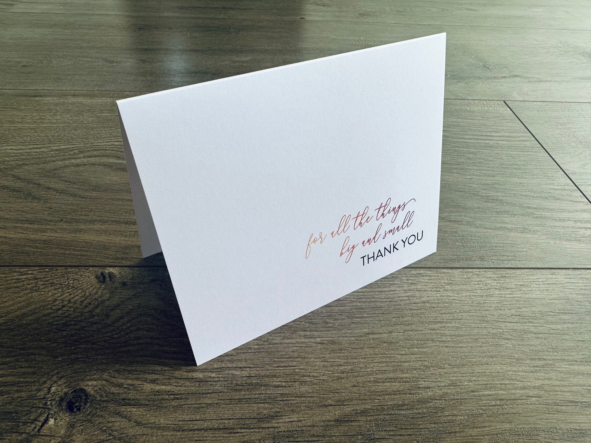 A folded white notecard is propped up on a wooden floor. The front of the card reads: for all the things, big and small, thank you