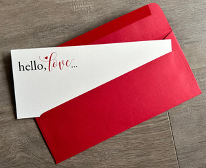 A textured white notecard is angled out of a bold red #10 envelope. Both lie flat on a gray wooden floor. The card reads, "hello, love" in the upper left hand corner. By Stationare.