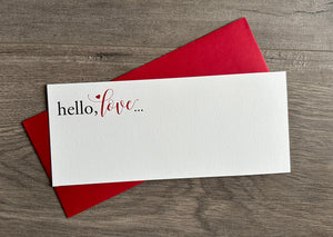 A narrow white notecard lies horizontally across a bold red #10 envelope. Both are lying flat on a gray, wooden floor. The card is of a white, textured stock and says "hello, love" in the upper left hand corner. By Stationare.