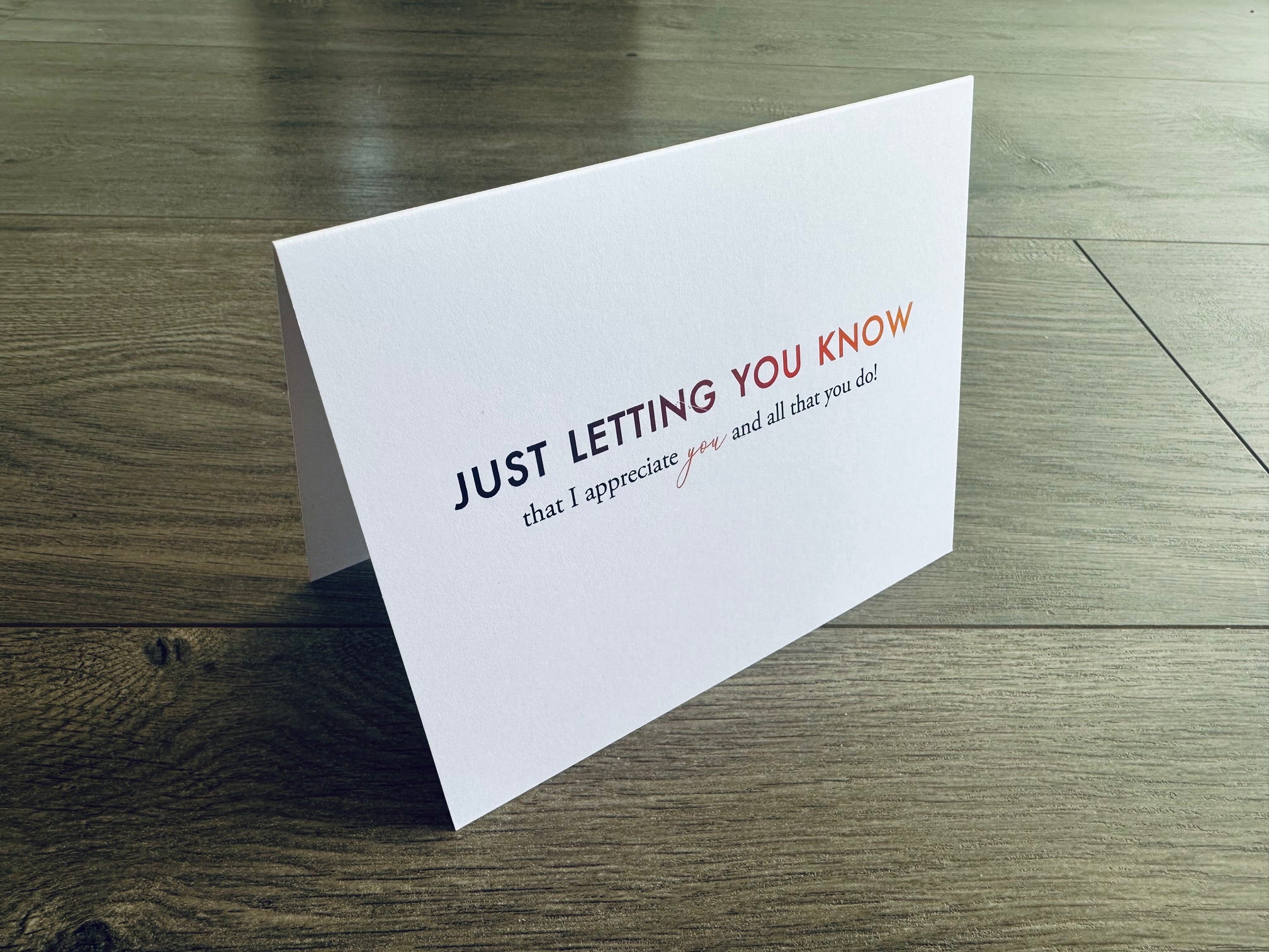 A folded white notecard is propped up on a wooden floor. The front of the card reads: just letting you know that I appreciate you and all that you do!