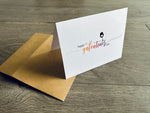 A kraft paper envelope is lying on a wooden floor with a small, white notecard folded and propped up on it. The card says, "Happy Galentine's Day" on the front with a small wine glass with a heart in it. by Stationare