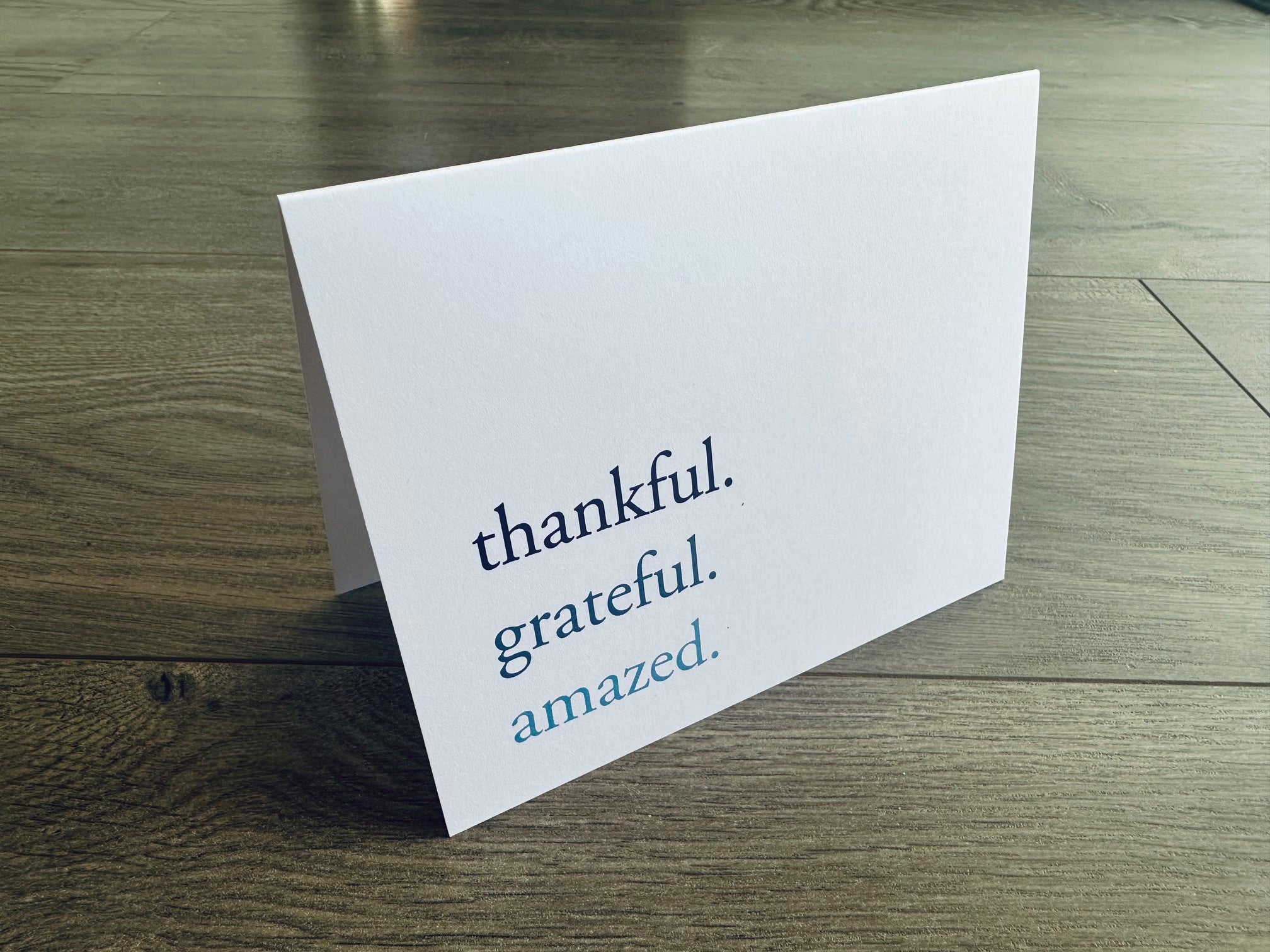 A folded white notecard is propped up on a wooden floor. The front of the card reads: thankful. grateful. amazed.