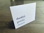 A folded white notecard is propped up on a wooden floor. The front of the card reads: thankful. grateful. amazed.