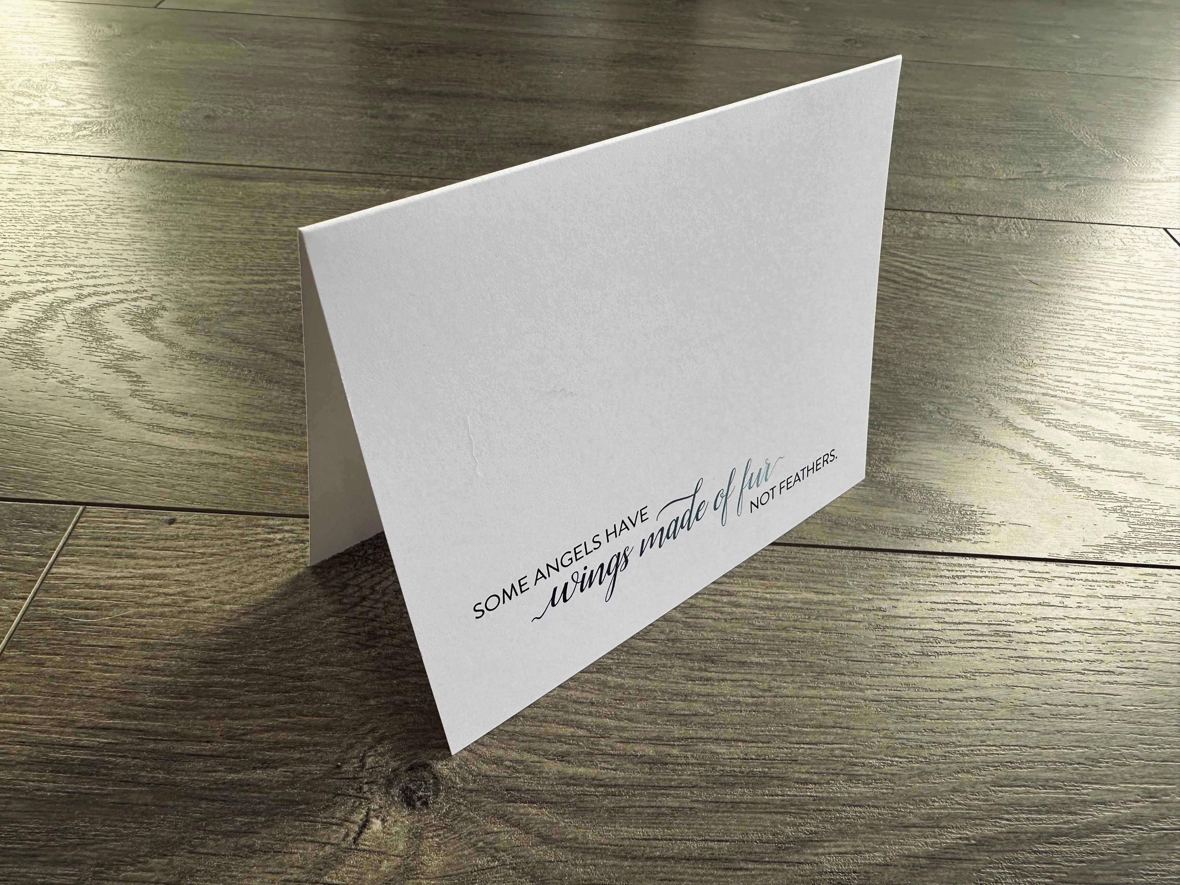 A white folded notecard is propped up on a wooden floor. On the front, it reads: "Some angels have wings of fur not feathers." Loss of Pet Collection by Stationare.