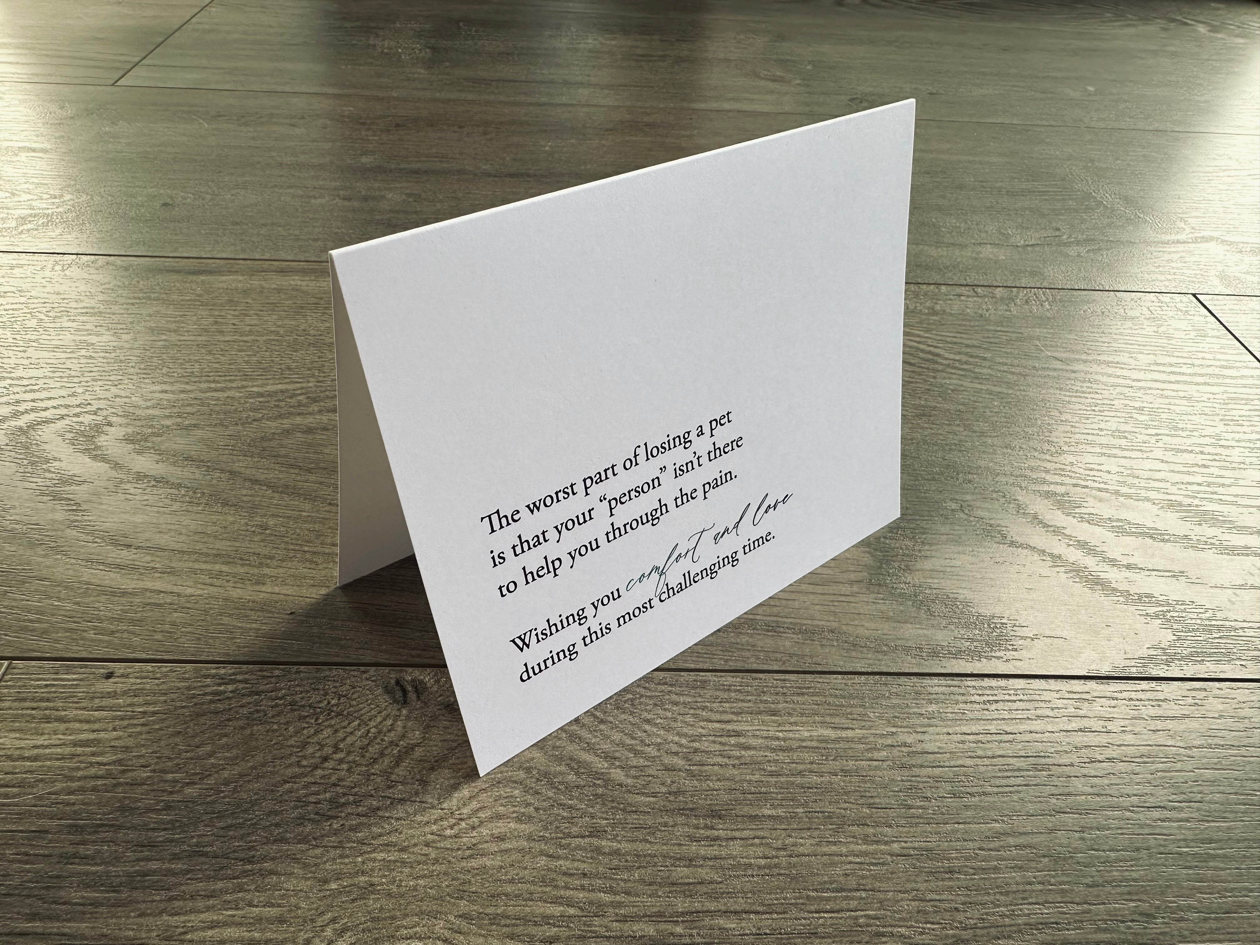 A folded white notecard is propped up on a wooden floor. It reads, "The worst part of losing a pet is that your 'person' isn't there to help you through the pain. Wishing you comfort and love during this most challenging time." Loss of Pet Collection by Stationare.