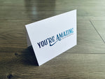 A folded white notecard is propped up on a wooden floor. The front of the card reads: You're amazing. Thank you so much!