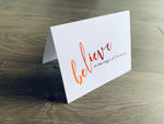 A white folded notecard is propped up on a wooden floor. The card says, "believe in the magic of Christmas." From the Meaning of Christmas Collection by Stationare.