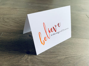 A white folded notecard is propped up on a wooden floor. The card says, "believe in the magic of Christmas." From the Meaning of Christmas Collection by Stationare.