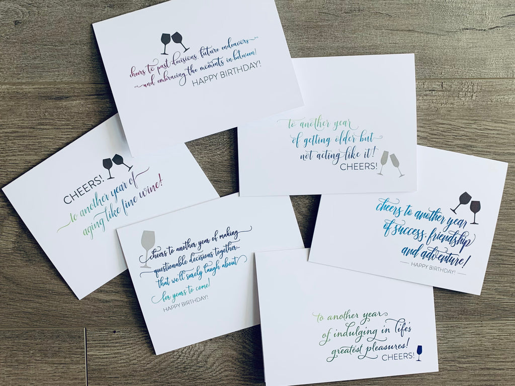 A collage of six white notecards are slightly overlapped and stacked on a wooden floor. Each has an array of blue, green and purple text with gray wine glasses. Birthday Cheers notecard collection by Stationare.