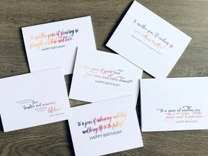 A collage of six overlapped white notecards lie on a wooden background. Each card has a birthday wish sentiment on it, written in an ombre of colors - orange, pink and purple. Birthday Wishes collection by Stationare