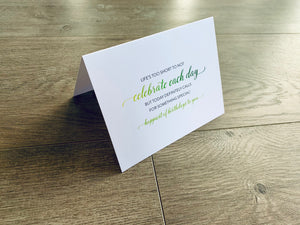 A white card is propped on a gray wood floor. The card reads, "Life's too short to not celebrate each day but today definitely calls for something special! happiest of birthdays to you" Birthday Inspirations by Stationare.