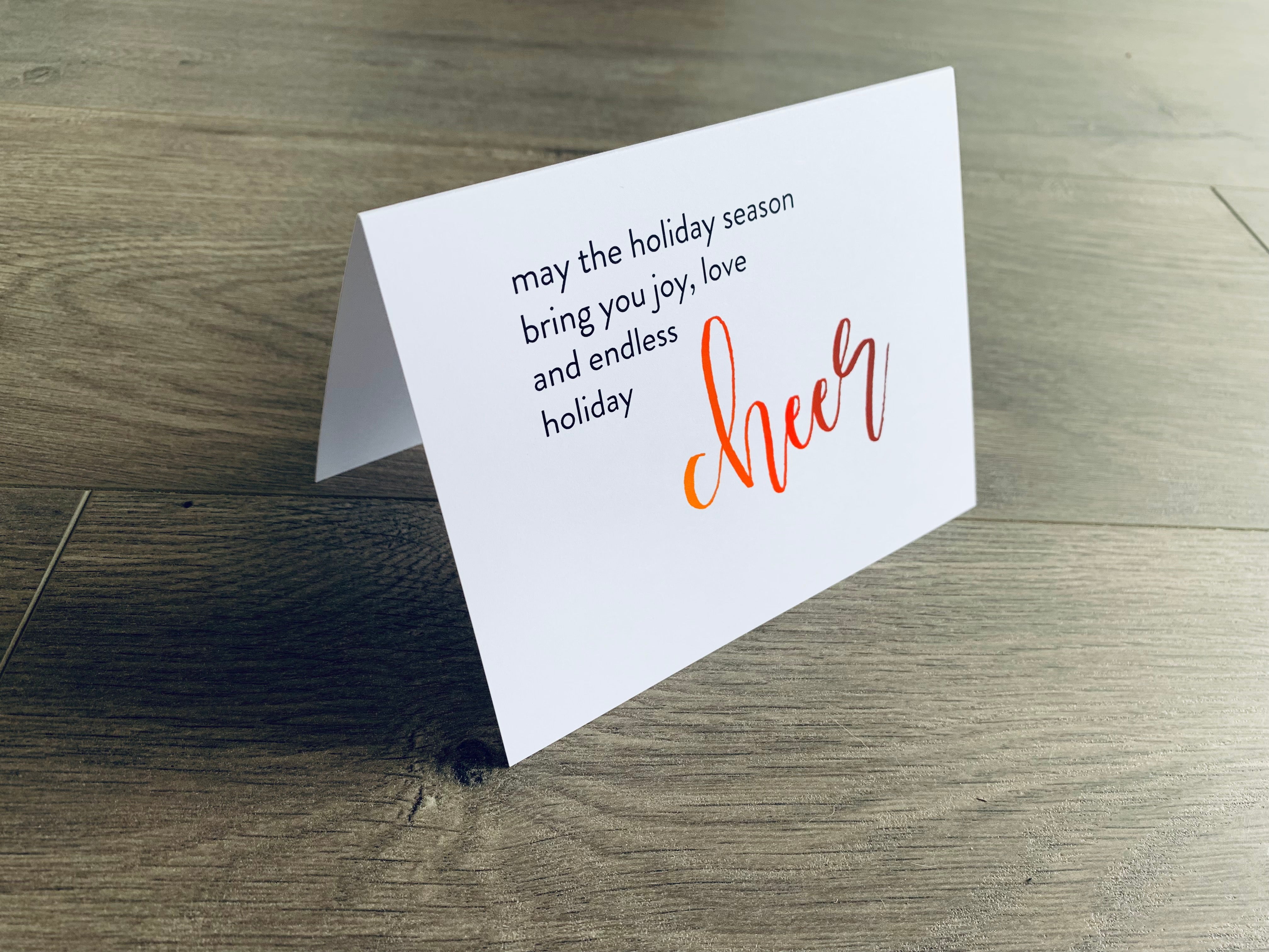 A white folded notecard is propped up on a wooden floor. The card says, "may the holiday season bring you joy, love and endless holiday cheer." From the Meaning of Christmas Collection by Stationare.