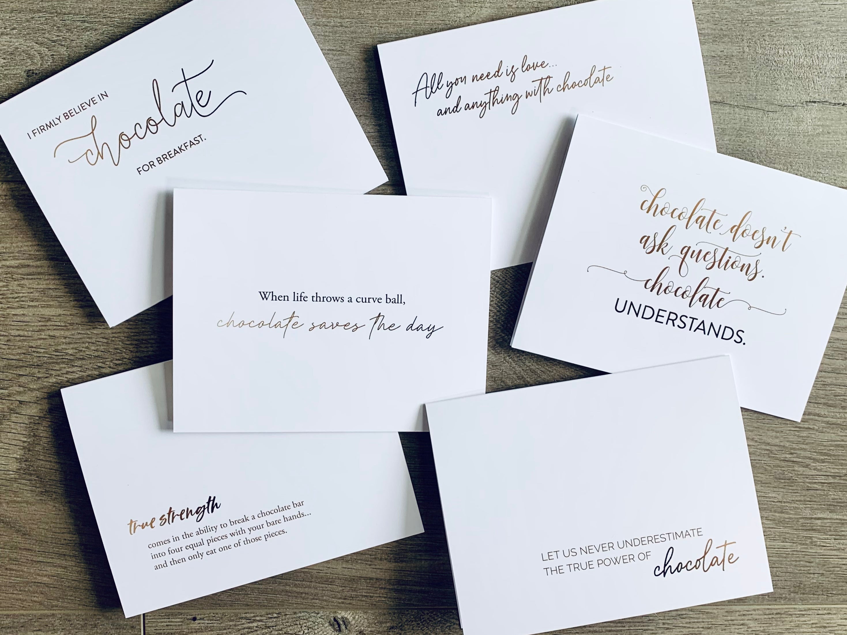 Six white notecards are lying on a wooden floor, overlapping one another. Each card has a chocolate-themed saying on the front in ranging brown tones. Chocolate Lovers Collection by Stationare.