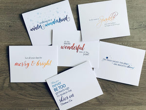 Six Christmas cards are overlapping each other on a wooden background. Each card has a traditional Christmas saying on the front. From Stationare's Christmas Magic Collection.
