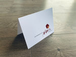 A white folded notecard is propped up on a gray wooden floor. The card reads, "I'm dreaming of a wine Christmas!" in a mix of sans serif and script fonts and a burgundy wine glass. Christmas Cheers by Stationare.