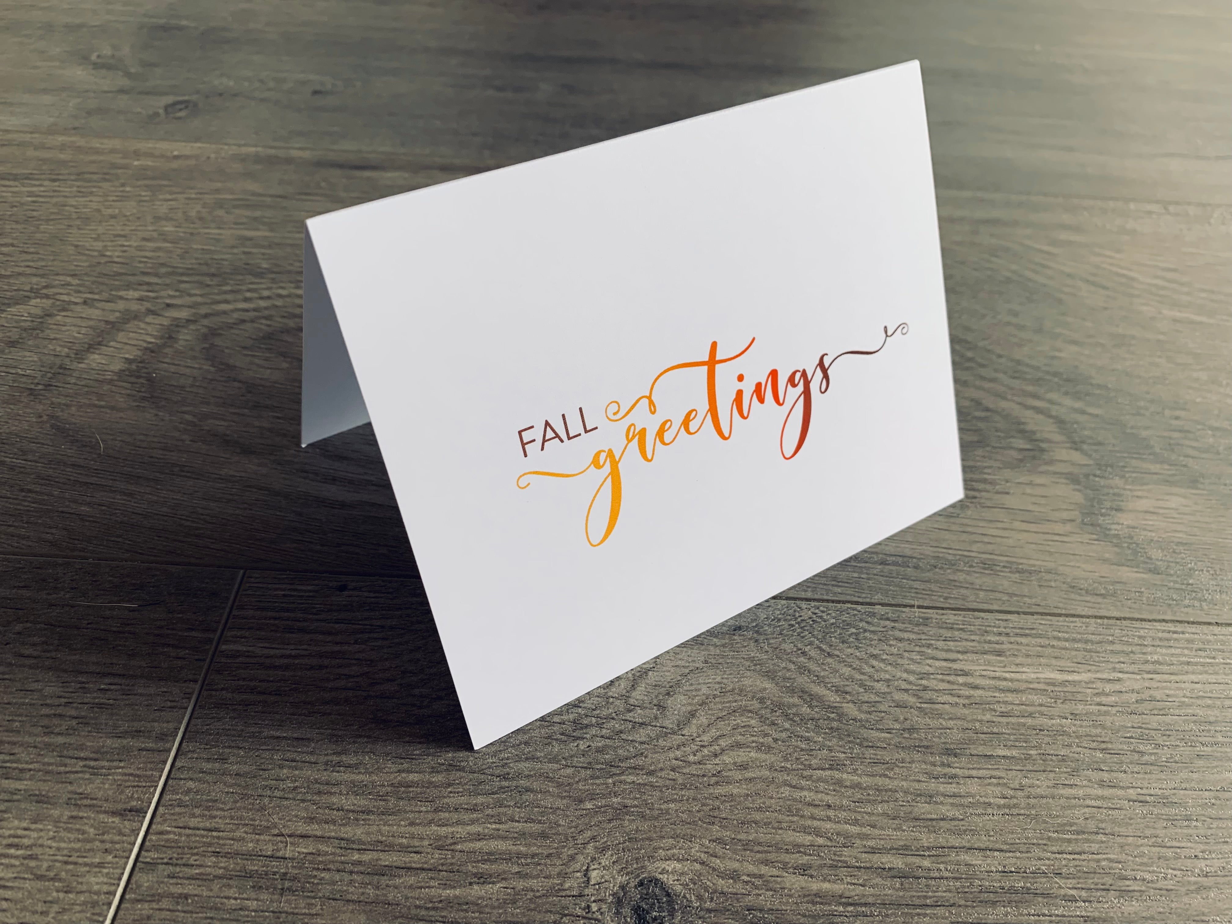 A white folded notecard is propped up on a wooden floor. The card says, "fall greetings." From the I Love Fall Collection by Stationare.