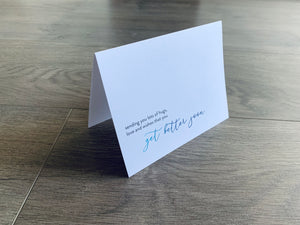 A white card with gray and blue text reads "Sending you lots of hugs, love and wishes that you get better soon."  From Stationare's Get Well Soon collection.