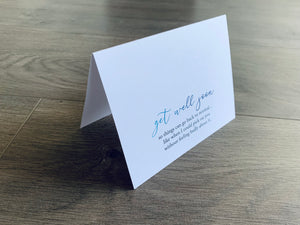 A folded white notecard sits on a wooden floor. It that, "Get well soon so things can go back to normal, like when I could pick on you without feeling badly about it." Joyful Well Wishes collection by Stationare.