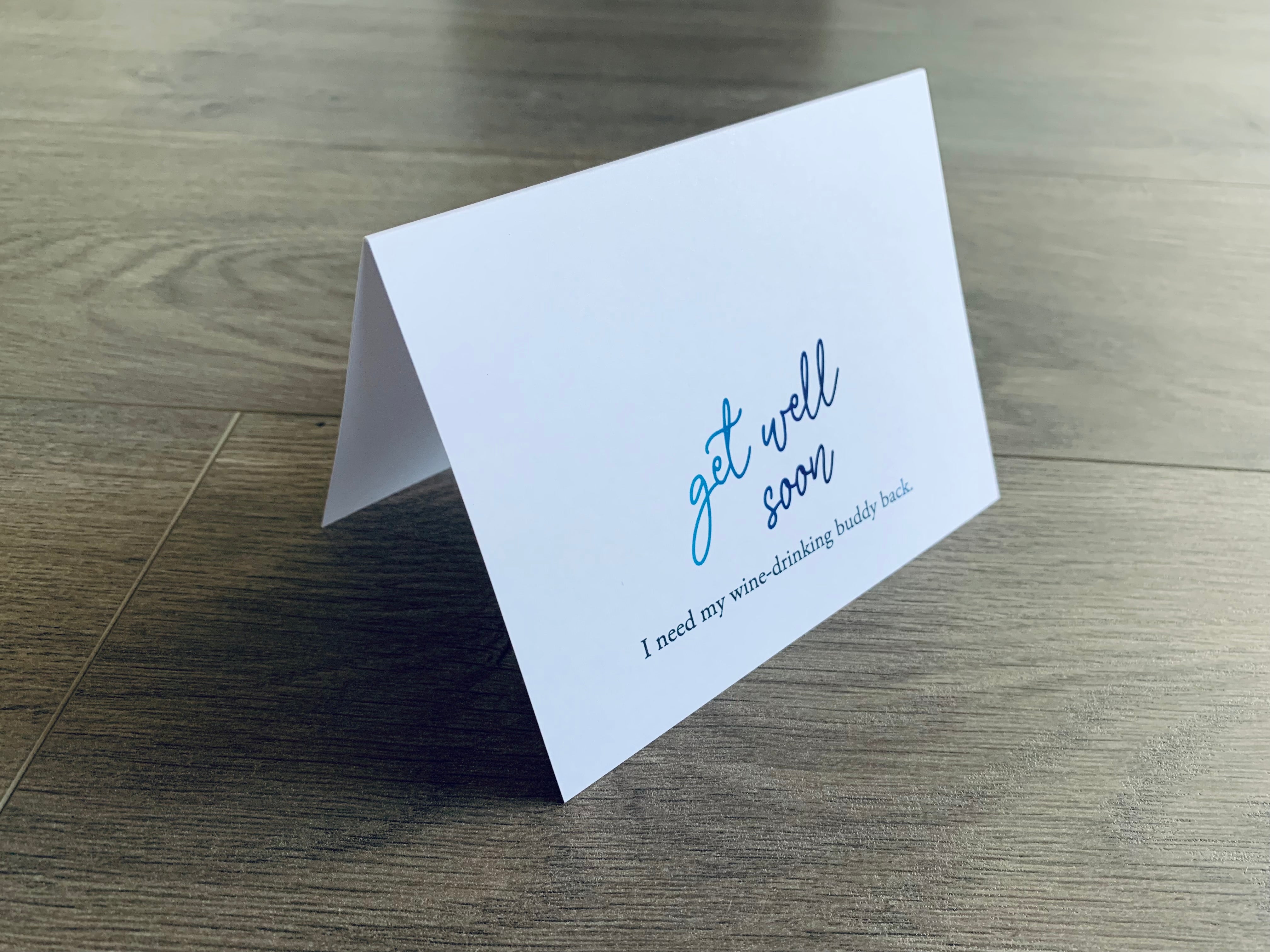 A white, folded get well soon card stands on a wooden floor and reads, "Get well soon. I need my wine-drinking buddy back." Joyful Well Wishes collection by Stationare.