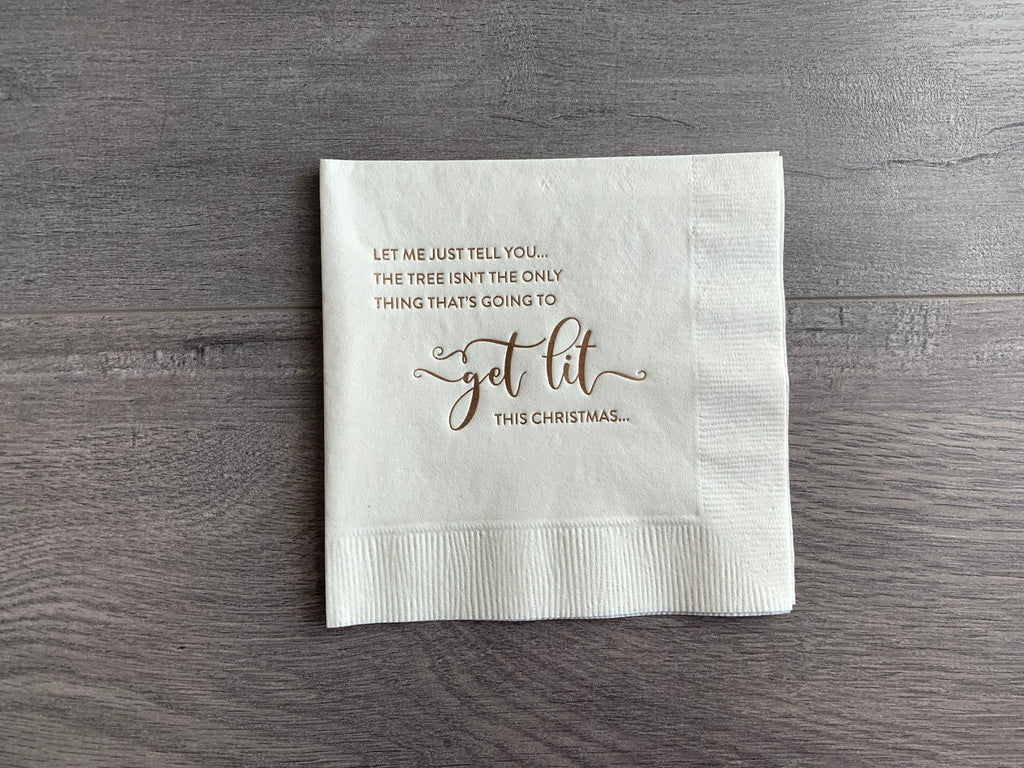 Off-white cocktail napkin lies on a gray wood background. In champagne foil it reads, "Let me just tell you, the tree isn't the only thing that's going to get lit this Christmas." 