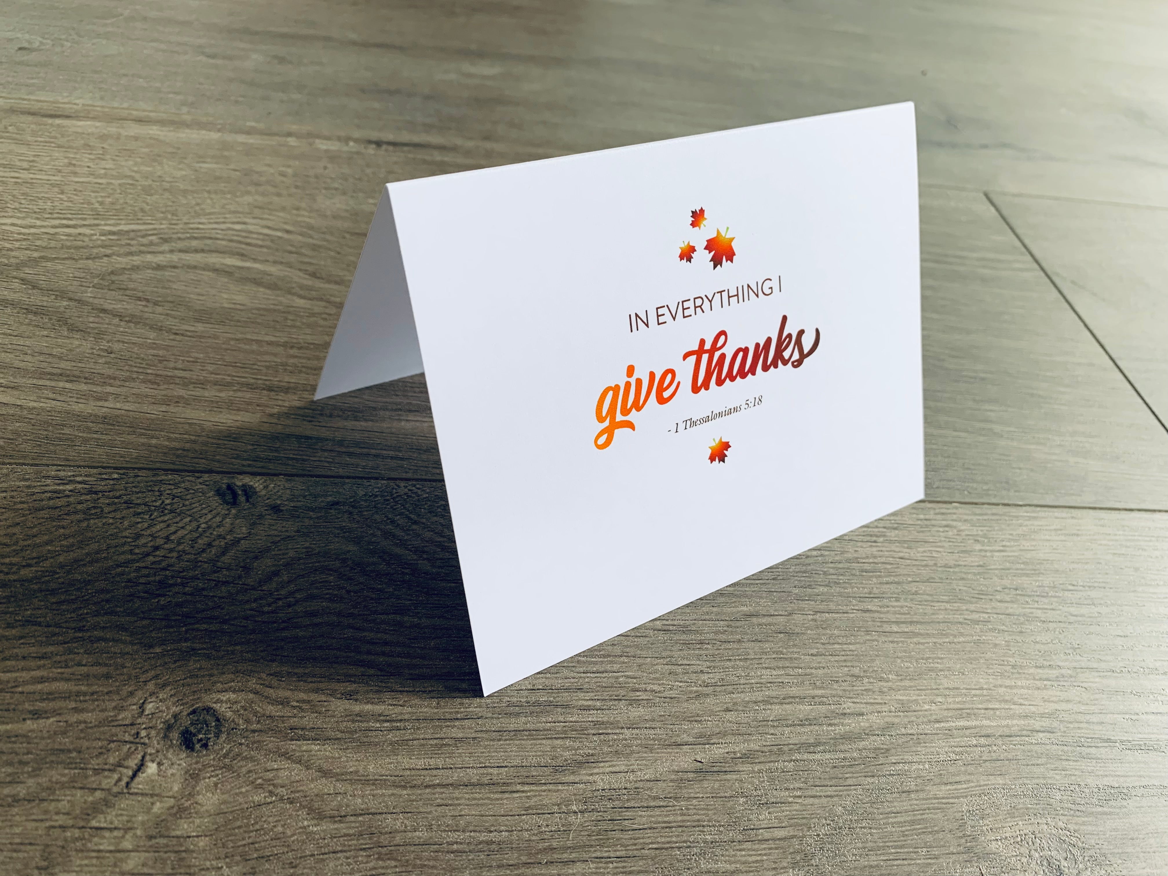 A white folded notecard is propped up on a wooden floor. The card says, "In everything, I give thanks" with small leaves around it. From the Giving Thanks Collection by Stationare.