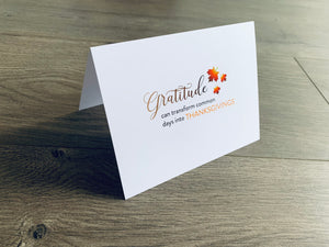 A white folded notecard is propped up on a wooden floor. The card says, "Gratitude can transform common days into Thanksgivings" with small leaves around it. From the Giving Thanks Collection by Stationare.