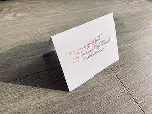 A white card is propped on a gray wood floor. The card reads, "to a year of great food, fine wine and extra dessert! happy birthday!" Birthday wishes collection by Stationare