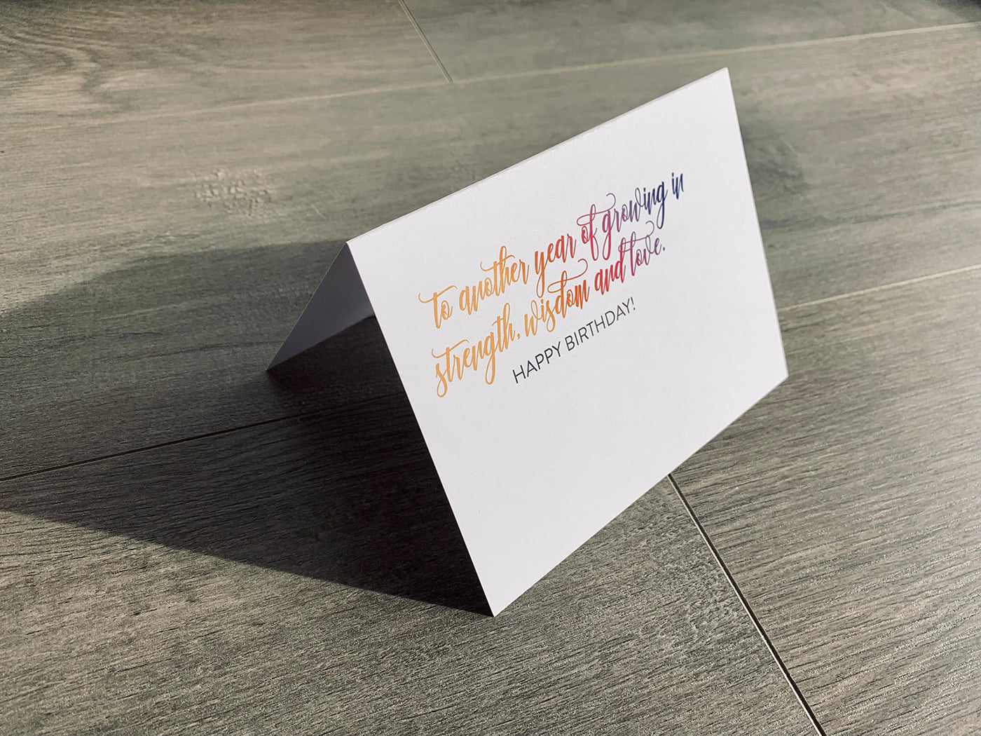 A white card is propped on a gray wood floor. The card reads, "to another year of growing in strength, wisdom and love. Happy birthday!" birthday card by Stationare