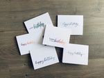 Six white notecards lie on a gray wood background. Each card says "happy birthday" in a script font in ranging ombre colors. Happy Birthday collection by Stationare.
