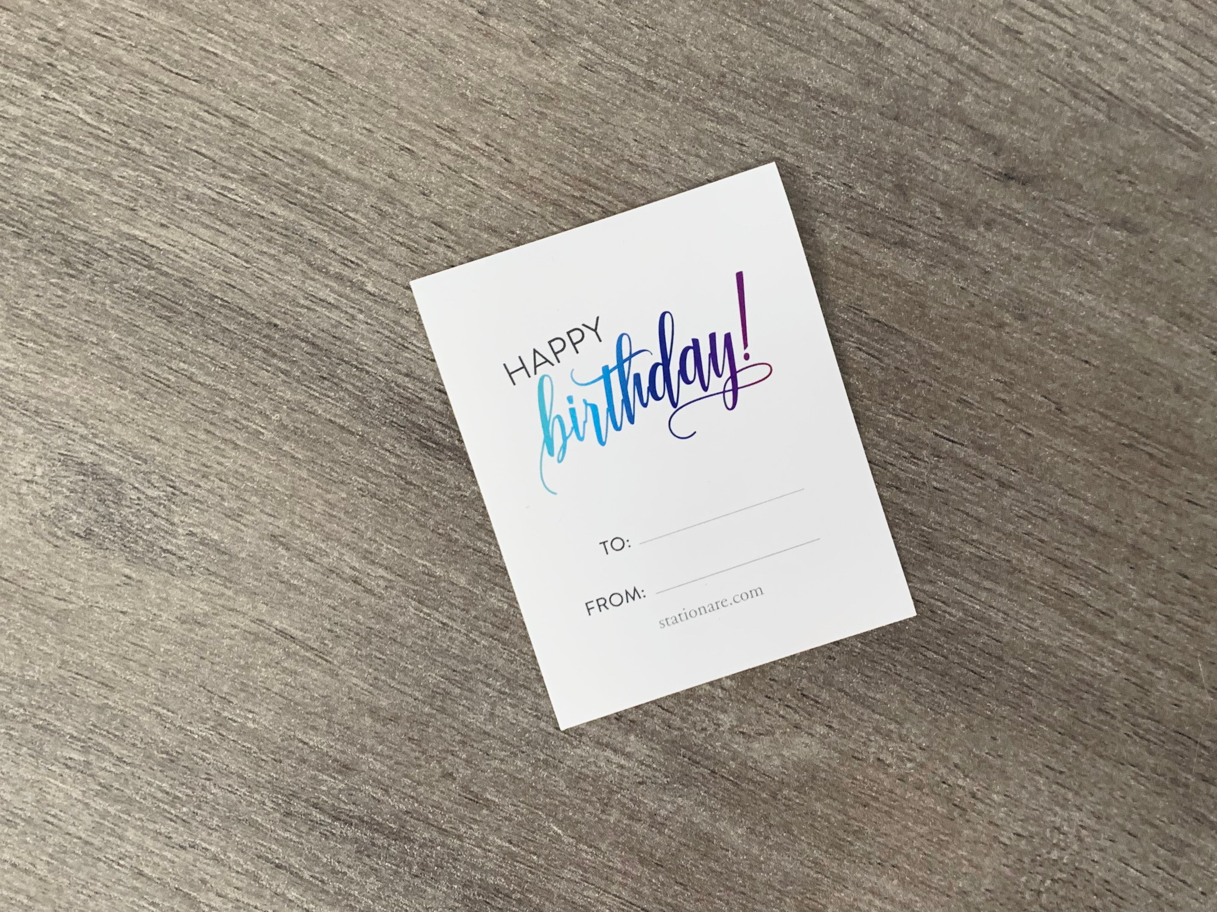 A white sticker gift tag rests on a gray wooden floor. The tag reads, "Happy birthday!" with a "to" and "from" area at the bottom. 