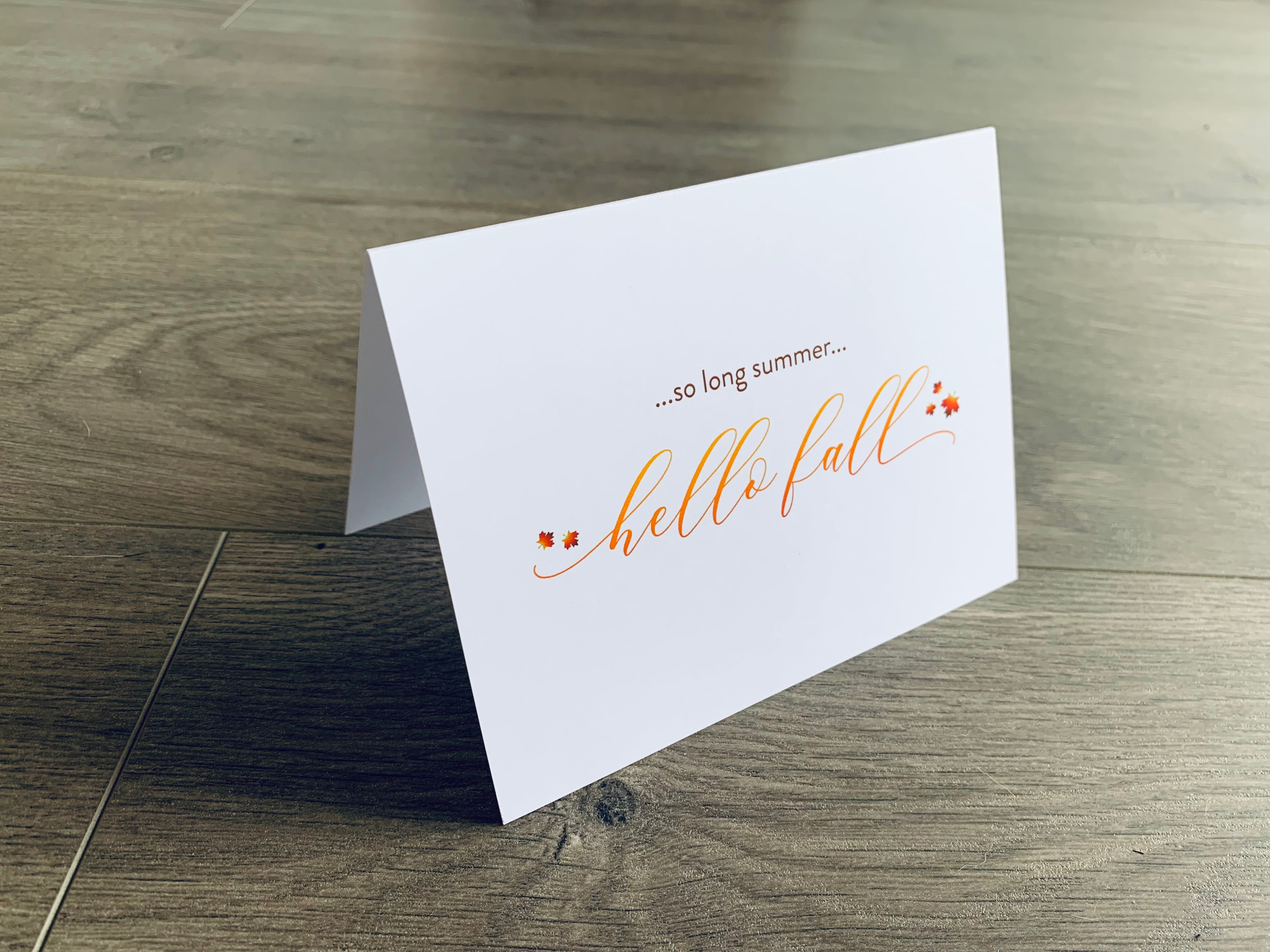 A white folded notecard is propped up on a wooden floor. The card says, "so long summer... hello fall" with small leaves around it. From the I Love Fall Collection by Stationare.