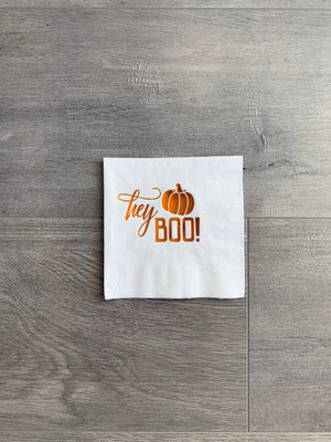 Straight-on view of a white napkin with shiny, orange copper foil printing. The printing reads, "hey, Boo!" with a small pumpkin above the word, Boo.