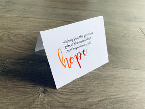 A white folded notecard is propped up on a wooden floor. The card says, "wishing you the greatest gifts of the season but most of all, hope." From the Meaning of Christmas Collection by Stationare.