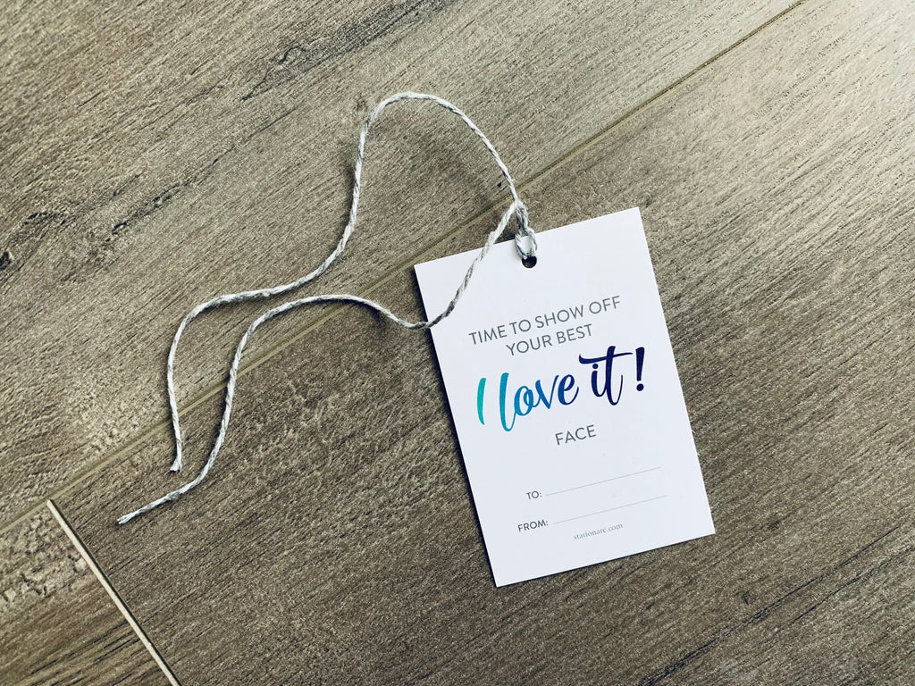 A white hanging gift tag with white and gray braided string lies on a wooden floor. The tag reads, "Time to show off your best I love it face" with a to and from section at the bottom. by Stationare