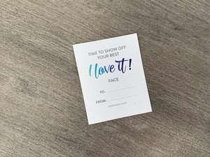 A white sticker gift tag lies on a gray wooden background. The tag says, "Time to show off your best I love it face." Underneath that saying is a To and From section to be filled in by the gifter.
