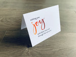 A white folded notecard is propped up on a wooden floor. The card says, "wishing you joy and cheer now and through the new year." From the Meaning of Christmas Collection by Stationare.