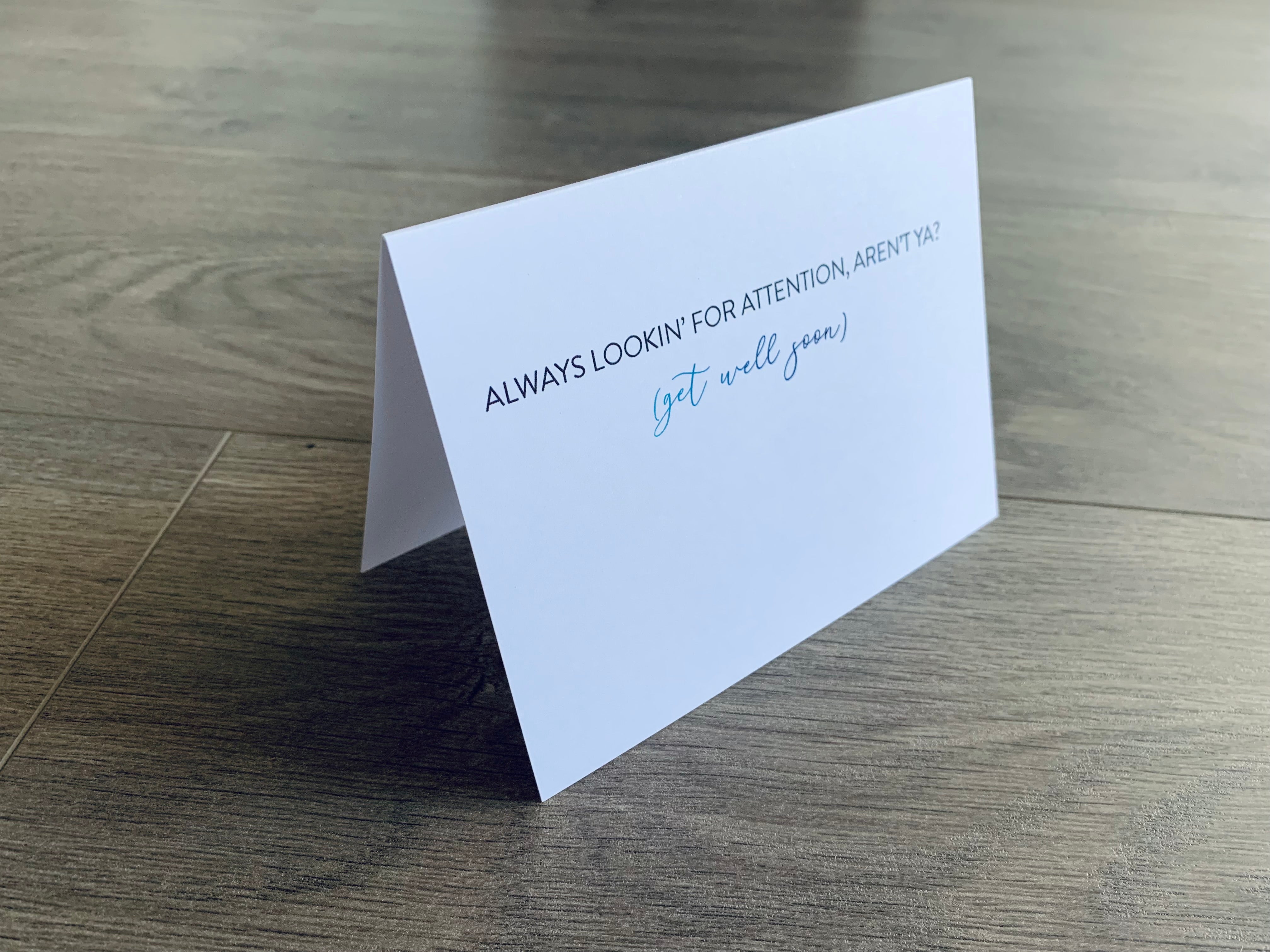 A white notecard sits up on a gray wooden floor that reads, "Always lookin' for attention, aren't ya?" In parens, it says "Get Well Soon." Joyful Well Wishes collection by Stationare.