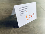 A white folded notecard is propped up on a wooden floor. The card says, "may your holiday season be filled with endless amounts of love." From the Meaning of Christmas Collection by Stationare.