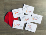 The six individual cards from Stationare's Meaning of Christmas collection are overlapped on a wooden floor, alongside a fanned-out stack of red envelopes.