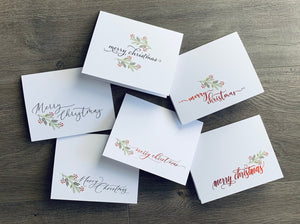 Six folded white Christmas cards lie on a gray wooden floor. Each card reads, "Merry Christmas," in a different script font and is accented with a small holly and branch design.